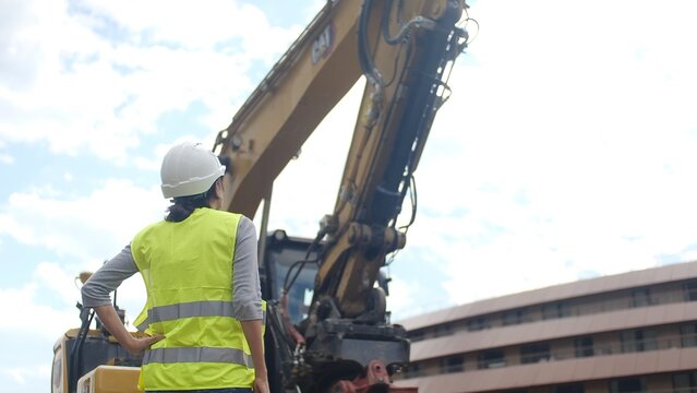 Woman construction worker in hard hat and safety vest standing at a building site, overseeing heavy machinery operation and project development, a symbol of women in stem