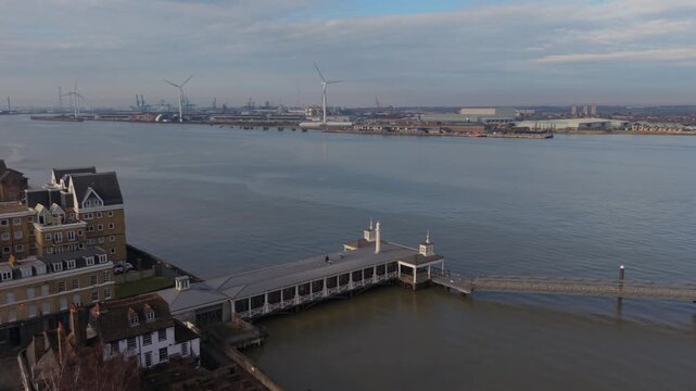 Historic Gravesend Pier on the river Thames. Aerial view flying towards and above the pier. Tall spinning wind turbines at Tilbury Docks as the industrial background. Sunny day with clouds in winter.