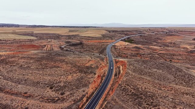 A road winds through a vast dry landscape with patches of brown and orange soil. Small towns can be seen in the distance. The sky is overcast, giving a gray look to the scene.