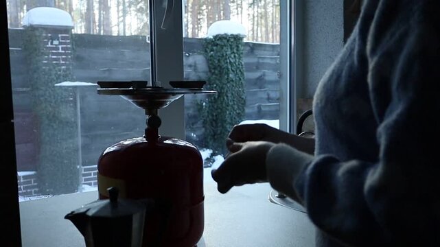 A young Ukrainian woman lights a gas stove with a match to make herself coffee during a power outage. The intimate kitchen scene highlights everyday resilience and adaptation to electricity shortages 