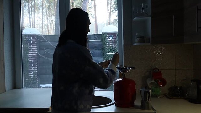 A young Ukrainian woman lights a gas stove with a match to make herself coffee during a power outage. The intimate kitchen scene highlights everyday resilience and adaptation to electricity shortages 