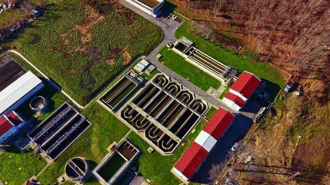 A view from above shows a water treatment facility with various pools and buildings arranged in a planned layout surrounded by green grass and brown trees.