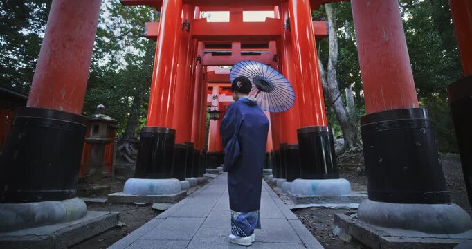 Woman in beautiful kimono and holding umbrella standing in front of ancient line of torii gates, slow motion