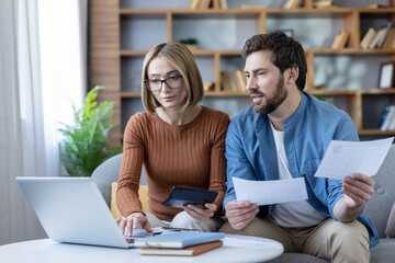 Couple sitting on a sofa in a home office, calculating their budget with a laptop and calculator, reviewing financial documents for taxes and planning future investments © Liubomir