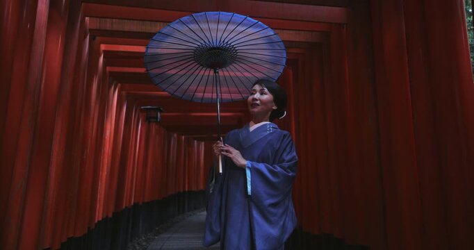 Cinematic slow motion of Japanese woman in kimono and holding umbrella under massive line of tori gates