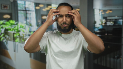Obraz premium Man pressing temples with both hands in a building restaurant interior, eyes closed and furrowed brow showing tension; frustration.