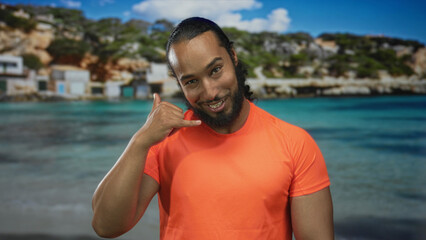Fototapeta premium Man pointing finger at camera on sandy beach by turquoise sea wearing bright orange shirt and beard; playful confidence.