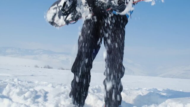 Snowboarder lifts snowboard from snow and shakes off stuck snow, cleaning board surface. Mountain winter sport, passion for snowboarding on frosty winter day.