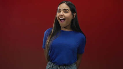 Fototapeta premium Woman with open mouth pointing right in a red studio wearing blue tshirt and jeans, long hair and hands behind back gesturing; surprise.