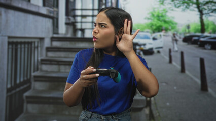 Young hispanic woman gamer holds a wireless gamepad and cups her ear with hand while listening intently on a street lined with buildings  curiosity engagement. © Krakenimages.com