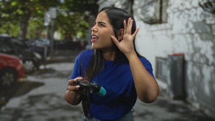 Woman holding gamepad cups her ear with raised hand and puzzled expression on a street next to parked cars and a concrete wall  curiosity challenge. © Krakenimages.com