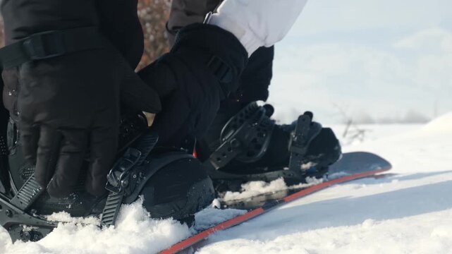 snowboarder hands in gloves fastening snowboard bindings on boots, tightening to proper tension. Rider standing on board securing bindings. Snowboarding as winter sport with extreme elements.