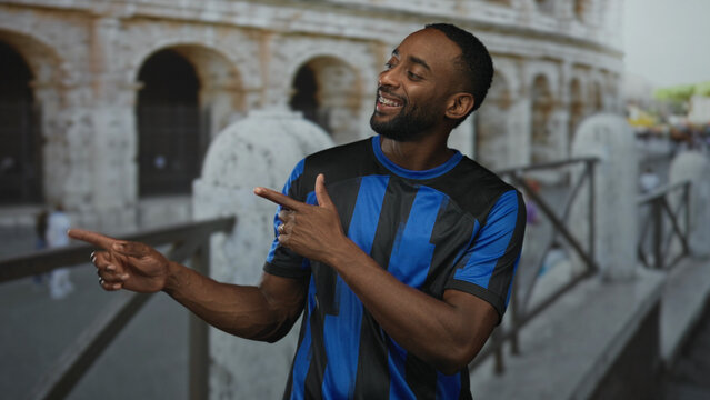 Man points finger with bare forearms wearing blue soccer jersey at roman colosseum building near ancient stone arches; team pride.