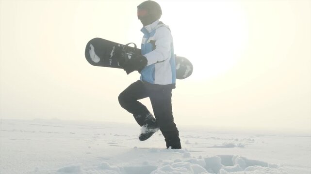 Slow motion confident steps of snowboarder in deep snow wearing helmet and ski goggles, carrying snowboard for mountain descent. Young rider aiming to beat personal record.
