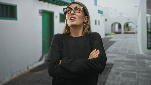 Young woman covering ears with hands and oversized glasses on a sunlit whitewashed street; frustration.