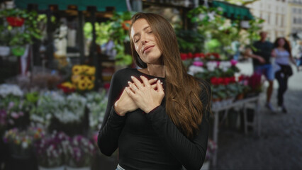 Fototapeta premium Woman holding hands over chest at a street flower market with closed eyes and long hair; serenity gratitude.