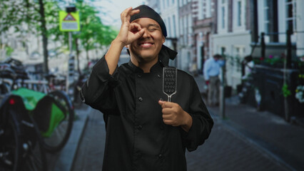 Hispanic man chef in black uniform holds metal spatula and makes ok sign toward camera on a busy street  street food joy. © Krakenimages.com
