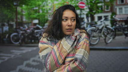 Fototapeta premium Young hispanic woman with hands clasped to mouth and nose, wearing patterned jacket, standing on a street with parked bicycles and brick townhouses; contemplation.