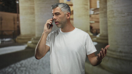 Man holding phone to ear with mouth open and pointing finger in building colonnade setting  fatigue distraction. © Krakenimages.com