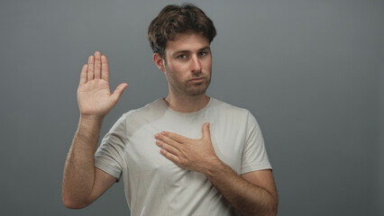 Man raising right palm and pressing left hand to chest in studio with gray wall; sincerity promise...