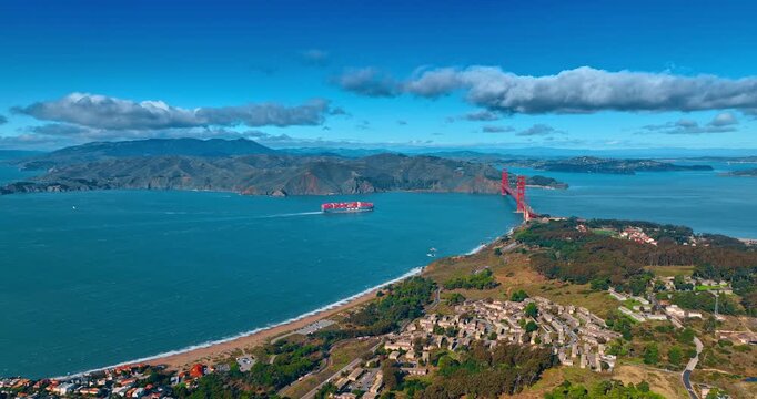 Large freight ship loaded with colorful containers moves slowly by the blue waterscape. Cargo vessel moves towards the Golden Gate Bridge in San Francisco, California, USA.