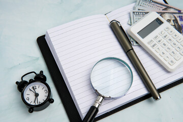 Open notebook with calculator, magnifying glass, alarm clock, pen, and US dollar bills, representing financial planning, budgeting analysis, money management, and accounting review.