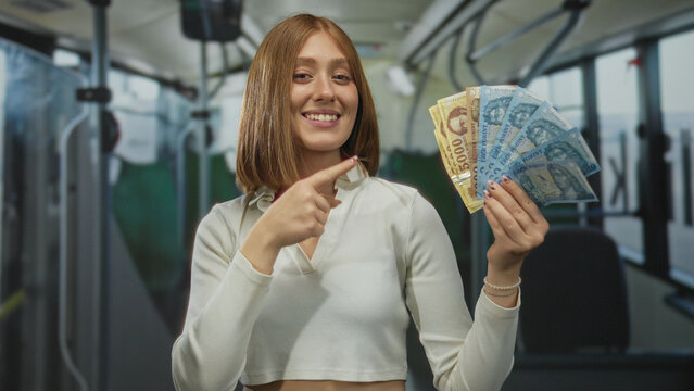 Young blonde woman on a bus showing hungarian forint banknotes while smiling indoors, highlighting public transport and currency.