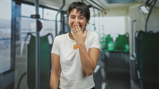 Woman with short hair covering mouth with hand, wearing rings and an orange pendant on a white t shirt inside a bus on street, smiling broadly; surprise.