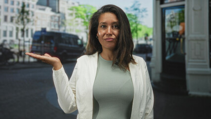 Fototapeta premium Woman with palm up hand gesture and white jacket, brunette middleaged subject standing near a van and building on a city street; bemusement indecision.