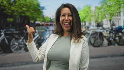 Fototapeta premium Brunette middle age woman smiling and giving a thumbs up while pointing at parked bicycles on a street lined with trees; confidence travel.
