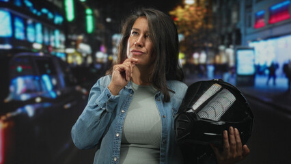 Fototapeta premium Woman middleaged brunette holding black motorcycle helmet, hand on chin in busy city street at night with neon signs and blurred pedestrians; thoughtful planning.