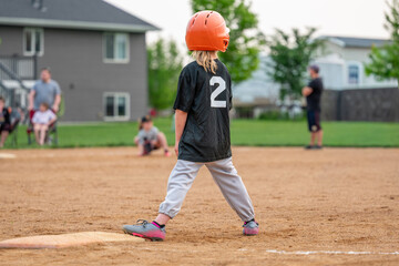 Child playing baseball on a field during a game in a neighborhood park in the evening with teammates and spectators present