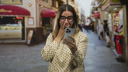 Fototapeta premium Woman covers mouth with hand while holding smartphone on a busy pedestrian street wearing glasses and patterned blouse; delight celebration.