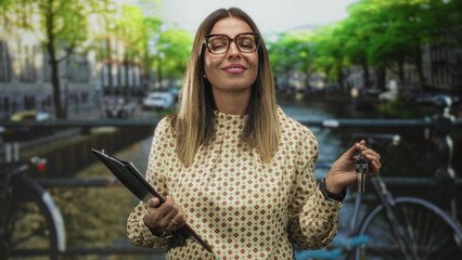 Hispanic woman real estate agent holding keys and clipboard, eyes closed and smiling while standing on street by canal and bikes; confidence home sale success.