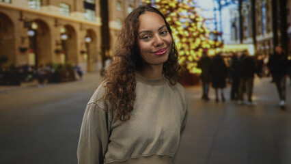 Obraz premium Woman smiles with visible face and slight head tilt on busy city street beside lit holiday tree and blurred crowd; joy confidence.