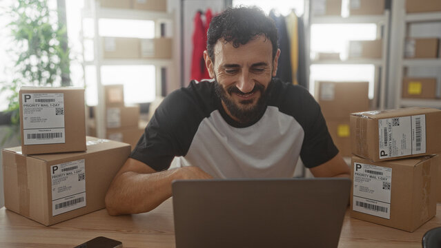 Man typing on laptop amid stacked packaging boxes and shipping labels in a building workspace for an ecommerce small business; entrepreneurial focused.