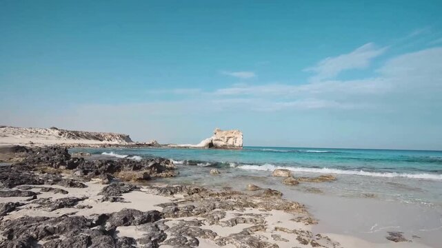 A serene tropical beach at morning light where gentle ocean waves wash over golden sand and rugged rocks along a coastline under a clear blue summer sky on cleopatra beach matrouh