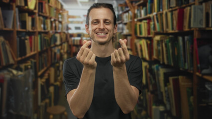 Man hispanic young long hair smiling and making finger heart gesture with bare fingers, wearing black t shirt, standing between bookshelves in library building  affection. © Krakenimages.com