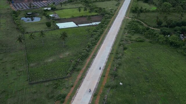 Aerial View Rural Highway Cutting Through Farmland, Long Straight Road Bordered By Green Fields And Scattered Farm Buildings, Palm Trees And Irrigation Ponds Visible, Minimal Traffic, Drone Survey
