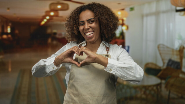 Young woman baker wearing apron in hotel lobby forms heart with hands while smiling broadly and looking at camera; joy hospitality welcome.