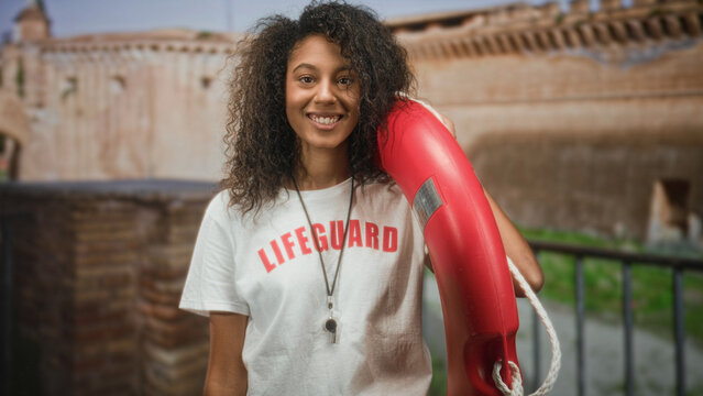 Teenage lifeguard woman holds red lifebuoy on shoulder and smiles wearing whistle necklace in front of historic building; confidence duty.