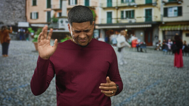 Man with beard and crewcut waving hands and grimacing on a cobblestone street in front of buildings and fountain; disgust.