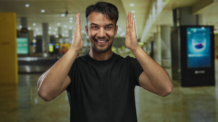Obraz premium Young hispanic man smiling inside airport terminal with hands framing face conveying joy and excitement in modern indoor travel environment.