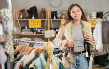 Positive young girl trying on fur coat while shopping in clothing store