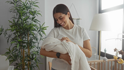 Woman smiling while cradling a swaddled baby beside a wooden crib and floor lamp in a bright nursery, holding the blanket close; maternal serenity.