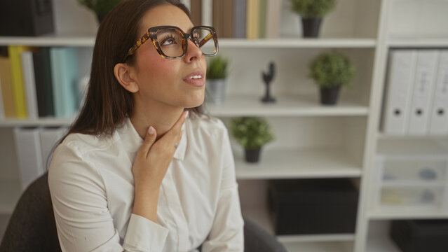 Woman holding throat with hand wearing tortoiseshell glasses and white blouse seated in office building with shelves and potted plants; discomfort illness.