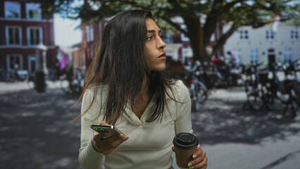 Fototapeta premium Young hispanic woman holding smartphone and coffee cup, squints eyes on cobblestone street with parked bicycles; urban frustration.