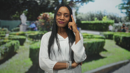 Black woman points finger to temple in a manicured garden wearing white blouse and braided hair;...