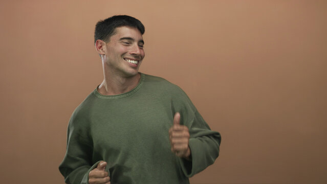 Man smiling and dancing with raised hands and thumbs up gesture in studio wearing green sweater; pure joy.