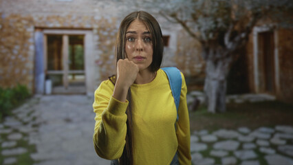 Fototapeta premium Young student woman in yellow sweater with blue backpack clenches fist near chin facing camera in front of stone building; defiance resilience.